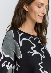 Czarny sweter z abstrakcyjnymi białymi i szarymi wzorami. Tkanina ma fakturę, luźny krój i szeroką okrągłą szyję.