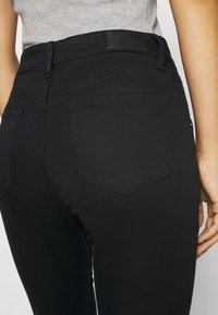 Primer plano de una persona que lleva jeans ajustados negros con un bolsillo trasero y una camiseta de canalé gris claro metida por dentro.