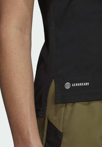 Débardeur noir sans manches avec une texture en maille et un logo, associé à un short vert olive. Le matériau semble léger et respirant.