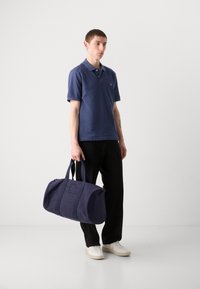 Navy-Dufflebag aus strukturiertem Stoff, mit doppelten Griffen, Logo-Stickerei auf der Vorderseite und runder Form. Wird von einer stehenden Person getragen.