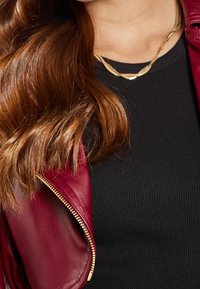 Giacca in pelle bordeaux con dettaglio zipper, abbinata a un top nero a coste e collane d'oro sovrapposte, con capelli castani mossi.