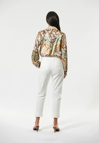 Blouse en soie avec des tourbillons colorés dans des tons de marron, turquoise et crème, associée à un pantalon blanc ajusté et des sandales à talons ouverts.