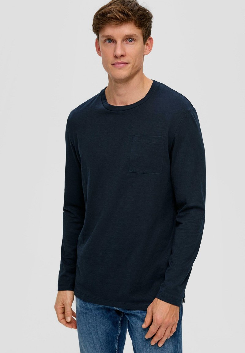 Granatowa koszulka z długim rękawem, uszyta z miękkiego materiału, z okrągłym dekoltem, z kieszonką na lewej piersi i bocznymi rozcięciami, zestawiona z niebieskimi dżinsami.