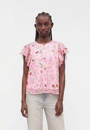 Frau mit geflochtenem Haar, die eine rosa Bluse mit Blumendruck und Rüschen an den kurzen Ärmeln sowie hellbeige Hose trägt, steht vor einem schlichten Hintergrund.