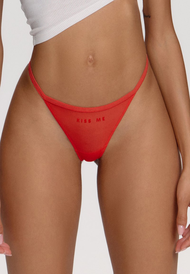 String rouge transparent avec le texte « EMBRASSE-MOI » imprimé à l'avant. Présente une couverture minimale, des bretelles fines et une texture lisse.