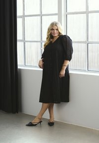 Robe noire longueur genou avec un décolleté en V, une fermeture à boutons et des manches bouffantes présentant un motif en chevrons. Associée à des escarpins noirs à bout pointu.