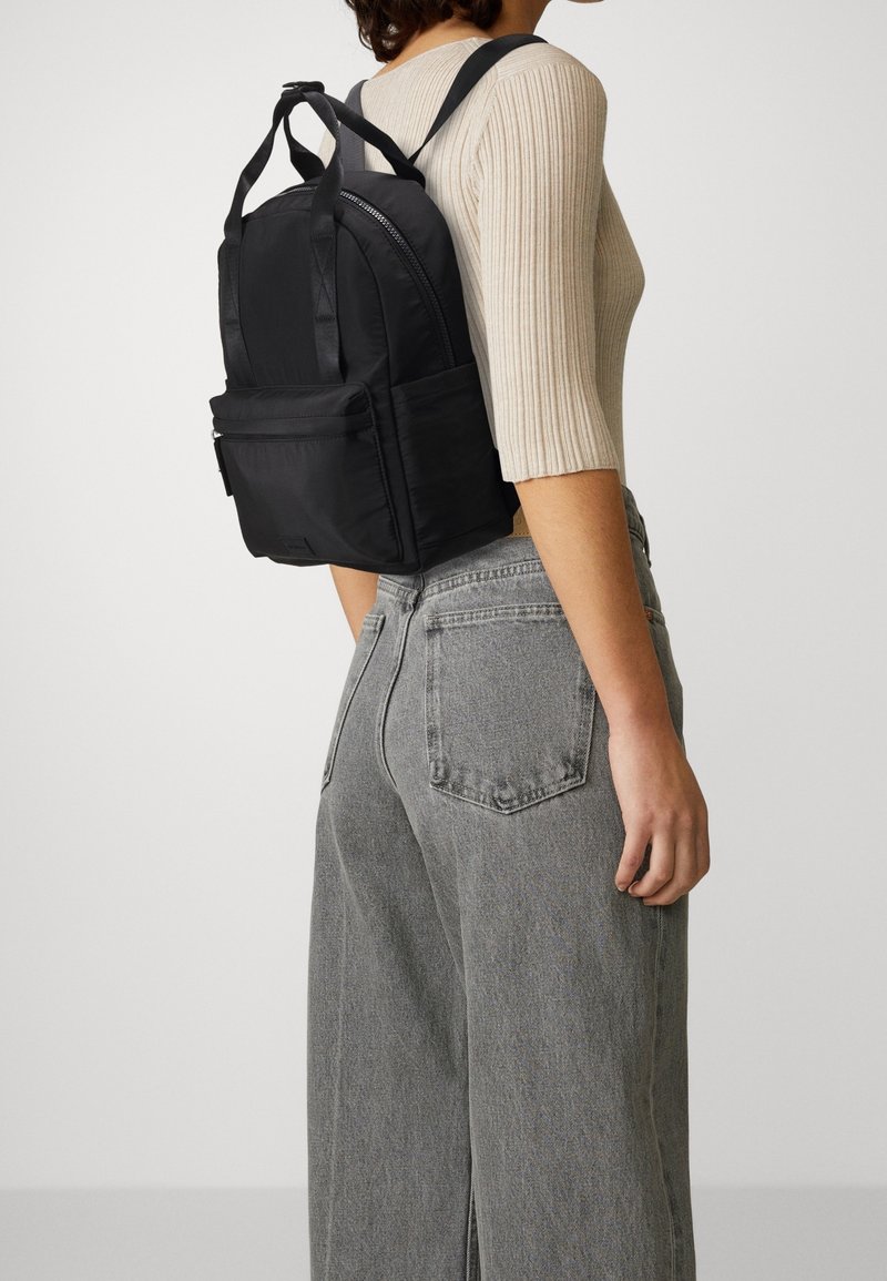 Personne portant un haut beige côtelé et un jean gris porte un petit sac à dos noir avec une poche avant et des poignées supérieures.