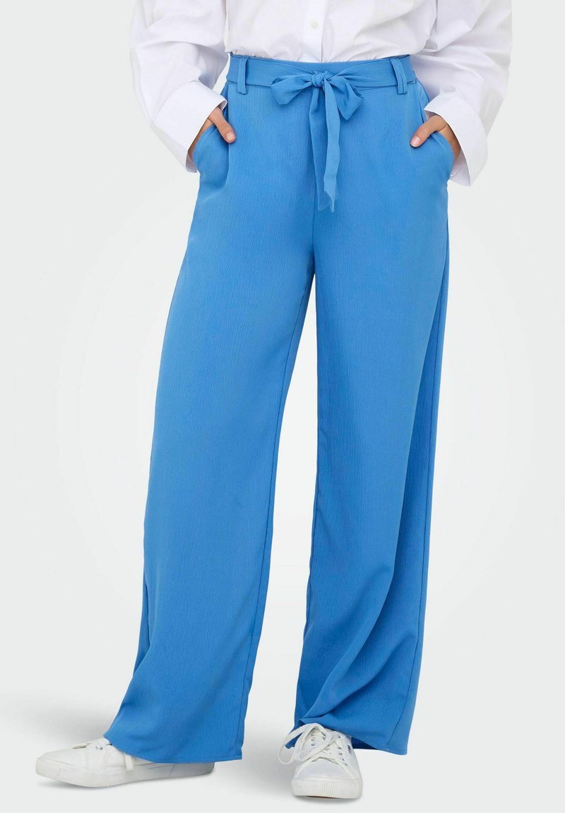 Pantalon à jambes larges bleu clair en tissu lisse, avec une taille nouée et des poches latérales, porté avec des baskets blanches.