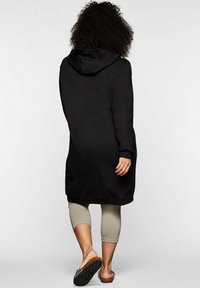 Musta huppu-sweatshirt-mekko, jossa on rento istuvuus, teksturoitu kangas, joustavat hihansuut ja pyöristetty helma. Yhdistetty vaalean värisiin legginsseihin ja liukumakiin sandaaleihin.