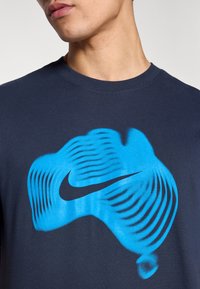 Μπλουζάκι από βαμβάκι σε ναυτικό μπλε χρώμα με μεγάλο, κυματιστό, μπλε γραφικό σχέδιο και μαύρο λογότυπο Nike Swoosh στο κέντρο. Στρογγυλή λαιμόκοψη, κοντά μανίκια.