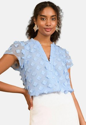 Blusa azzurra chiara a maniche corte con applicazioni floreali testurizzate e chiusura a bottoni frontale. Presenta un collo a scallop e una delicata rifinitura.