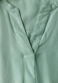 Blouse vert clair avec un col en V et une texture douce, dotée de coutures nettes le long des bords et d'un tissu lisse et légèrement brillant.