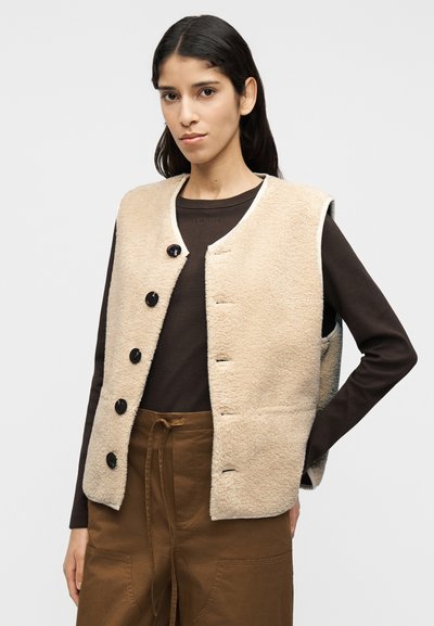 Μπεζ φλις γιλέκο με στρογγυλή λαιμόκοψη, μαύρα κουμπώματα και πλαϊνές τσέπες, φορεμένο πάνω από μακρυμάνικο τοπ σε σκούρο καφέ χρώμα.