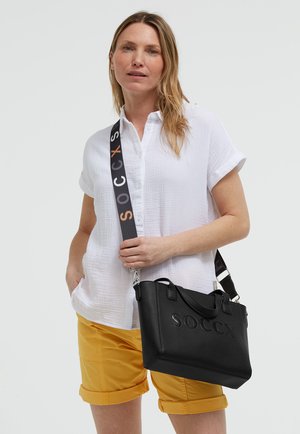 Frau trägt weißes kurzärmeliges Hemd und gelbe Shorts, trägt schwarze SOCCX Tragetasche mit gebrandetem Träger über einer Schulter.