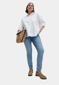 Bílá košile na knoflíky, světle modré džíny, hnědé mokasíny, držící texturovanou slaměnou psaníčko s černými akcenty, vystavené na bílém pozadí.