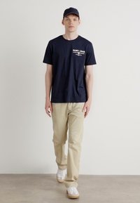 Middengrote navyblauwe katoenen t-shirt met een kleine witte print, gecombineerd met lichtbeige broek en witte sneakers. Het model draagt ook een bijpassende navyblauwe pet.