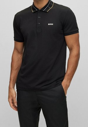 Mann trägt ein schwarzes kurzärmeliges Polohemd mit "BOSS"-Logo auf der Brust und am Kragen, kombiniert mit einer schwarzen Hose, steht mit einer Hand in der Tasche.