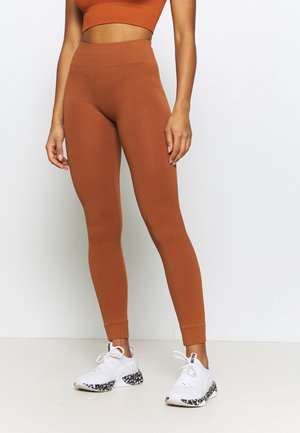 Femme portant des leggings côtelés rouille taille haute et un crop top assorti, avec des baskets Puma blanches aux semelles noires à motifs.