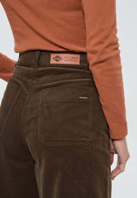 Pantalons en velours côtelé marron avec poches arrière et une étiquette en cuir comportant les détails de la marque. Le haut est une chemise à manches longues orange.
