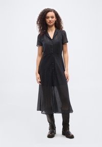 Γυναίκα στέκεται φορώντας μαύρο διαφανές midi φόρεμα με πουά, κοντά μανίκια και μαύρες μπότες μέχρι το γόνατο, μπροστά σε απλό ανοιχτό φόντο.