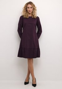 Purppurapituilla hihoilla varustettu mekko, jossa on pleatattu helma. Sujuva kangas, suora siluetti, pyöreä pääntie ja minimalistinen muotoilu.