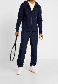 Laivastonsininen vetoketjullinen huppari, jossa viininpunainen vuoraus hupussa, yhdistetty samanvärisiin laivastonsinisiin collegehousuihin. Valkoiset tennarit oranssein yksityiskohdin. Pitelee tennismailaa ja palloa.