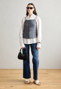 Gray linen top with thin straps over a white long-sleeve shirt, blue jeans, and cream slide sandals. Holding a small black handbag.