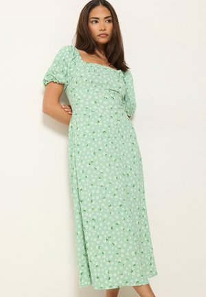 Donna che indossa un vestito verde chiaro a maniche corte con stampa floreale bianca, in piedi contro un muro chiaro e semplice, con le mani dietro la schiena.