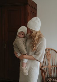Tuhkanvärinen neulemekko ja kermaiset pörröiset hatut, lapsella beige fleece-takki, taustalla puinen tuoli, lämmin sisävalaistus.