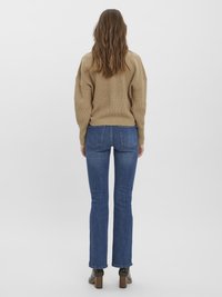 Žena otočená chrbtom, oblečená v béžovom pletenom svetri, modrých rovnomerných džínsoch a tmavých podpätkových členkových čižmách na bielom pozadí.