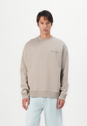 Sweatshirt beige chiaro con scollo rotondo, polsini e fondo a coste. Presenta un piccolo logo testuale sul petto. Indossato con jeans azzurri chiari.