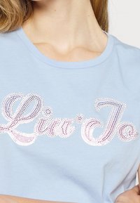 Mujer vistiendo una camisa azul claro con texto brillante de strass "Liu Jo" en el pecho, mostrando el escote y parte del brazo.