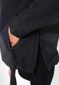 Black windbreaker jacket with a smooth texture, featuring zipped pockets and elasticated hems for adjustable fit.