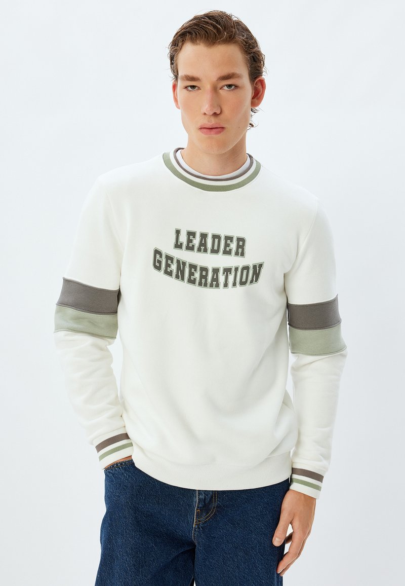 Vit sweatshirt med "LEADER GENERATION" i gröna bokstäver. Har grön- och grårandiga muddarn, rund halskrage och avslappnad passform.