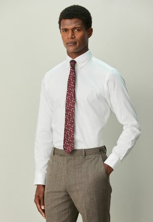 Mann trägt weißes Hemd mit Knöpfen, gemusterten kastanienbraunen Krawatte und braune Tweedhose, Hand in der Tasche, vor neutralem Hintergrund.