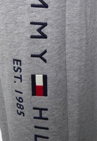 Calças de treino cinzentas com texto bordado em azul marinho "TOMMY HILFIGER EST. 1985" e um pequeno emblema em vermelho, branco e azul. Textura suave.