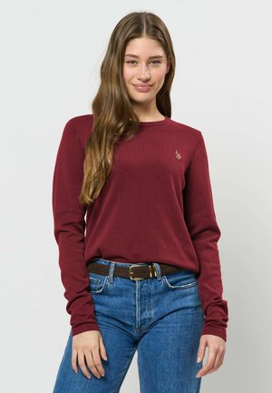 Jeune femme aux longs cheveux bruns, portant un pull bordeaux à manches longues et un jean bleu, debout devant un fond clair.