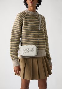 Maglione a righe in beige e oliva, con collo a coste. Borsa a tracolla grigia chiara con superficie testurizzata e scritta in rilievo. Gonna plissettata.
