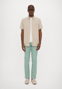 Camisa de botones beige de manga corta, camiseta blanca debajo, pantalones verde menta, zapatos beige y gorra marrón con acento de logo.