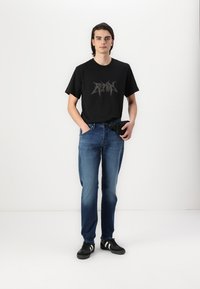 T-shirt en coton noir avec impression graphique argentée, associé à un jean en denim bleu. Les accessoires comprennent une ceinture noire et des baskets noires.
