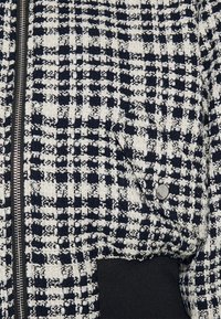 Veste en tweed présentant un motif à carreaux bleu marine et blanc, avec une fermeture éclair et un accent en tissu noir sur le côté. Finition texturée.