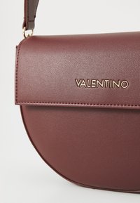 Bordeaux lederen crossbody tas met een ronde vorm, voorzien van een flap sluiting, gouden hardware en geperste "Valentino" logo.