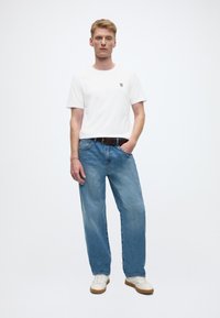 Bijela kratka majica s logotipom, uparena s svijetloplavim trapericama širokih nogavica i bijelim tenisicama s smeđim đonovima.