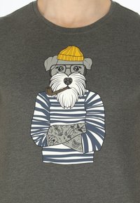 Grå T-shirt med en illustrerad hund som bär en gul mössa och glasögon, en randig tröja och tatueringsliknande mönster på sina armar.