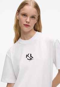 Jovem mulher com cabelo loiro longo, vestindo uma t-shirt branca com um logotipo preto da Karl Lagerfeld Jeans em um fundo branco liso.