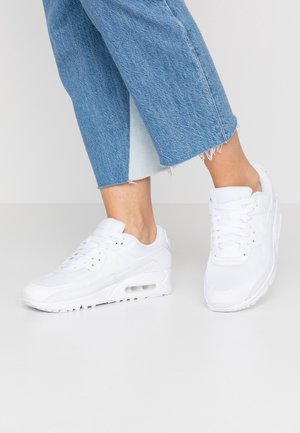 Osoba ubrana w krótkie niebieskie jeansy i białe buty Nike Air Max stoi na jasnoszarej podłodze.