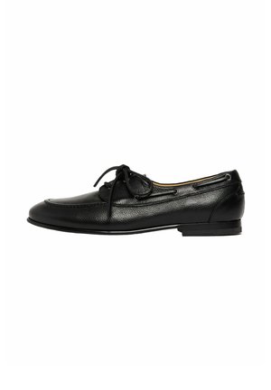 Mocasín de cuero negro con punta redondeada, detalles de costura, lazo de cordón en la parte superior y tacón bajo, mostrado en perfil lateral sobre fondo blanco.