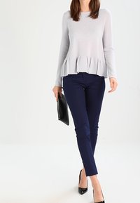 Lichtgrijze langemouwen sweater met een peplumzoom, gecombineerd met navy slim-fit broek en zwarte hoge hakken, met een zwarte clutch in de hand.