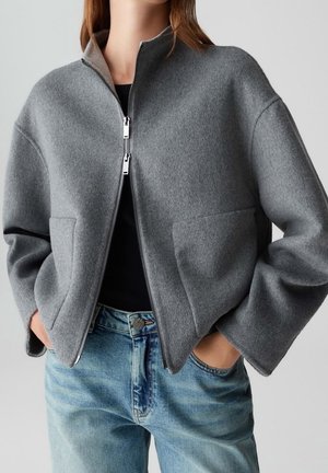 Mujer con chaqueta de lana gris con cremallera y grandes bolsillos delanteros y vaqueros azul claro, con las manos en los bolsillos, frente a un fondo liso.