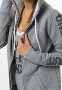 Sudadera gris con cremallera y cordones blancos, con bolsillos laterales y texto negro impreso en la manga. Llevada sobre una camiseta blanca en capas.